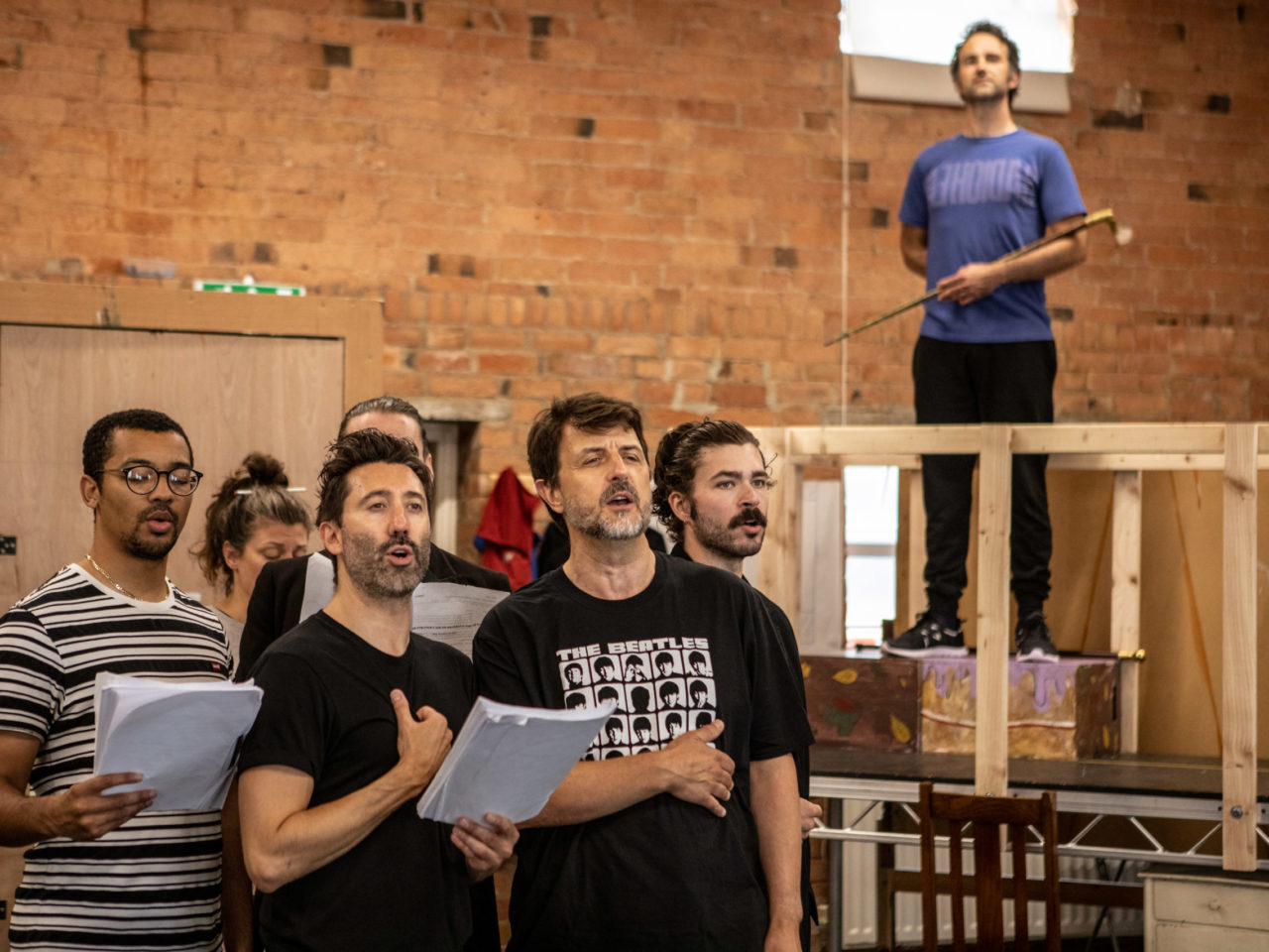 The male ensemble stood in the rehearsal rooms. Nick Shaw is in the background looming over the group as Mr Jekyll or Hyde.